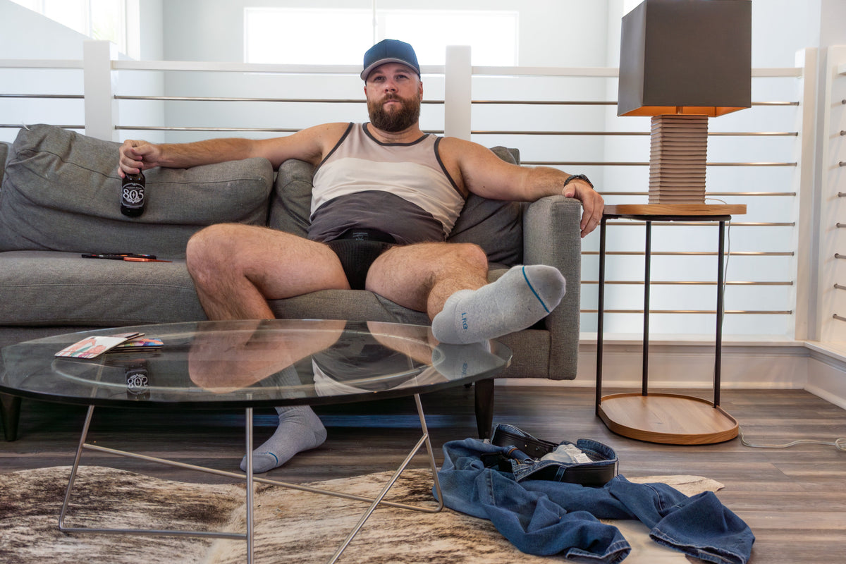 Scott Johnson relaxing on a gray sofa in a cap, striped tank and black jockstrap, holding a beer, with jeans on the floor.